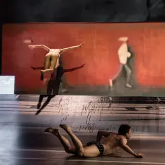 Andrzej Wencel w powietrzu, a na dole mężczyzna na scenie. Tło to malowidło z biegnącą postacią.