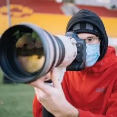 Fotograf sportowy w masce i czerwonej kurtce z aparatem Canon EOS-1D X Mark III czeka na akcję na tle trybun stadionu.