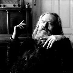 Miroslav Tichý, zamyślony, z długą brodą, siedzi w fotelu. W tle widać mebel z radiem i figurką.