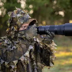 Osoba w kamuflażu liściastym z aparatem i długim obiektywem, gotowa do fotografowania ptaków w lesie.
