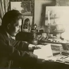 Tadeusz Rząca pochylony nad dokumentami przy biurku, w otoczeniu obrazów i lampy naftowej.