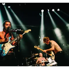 Energetyczna fotografia koncertowa: wokalista z tatuażami gra na niebieskiej gitarze, basista pochyla się nad swoim instrumentem, a perkusista wybija rytm.