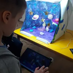 Chłopiec tworzy kosmiczną animację poklatkową. Na stole stoi diorama z rakietą, astronautą i planetami, a obok tablet z podglądem.