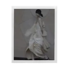 Modelka w zwiewnej, kremowej sukni i czarnym kapeluszu, w stylu Sarah Moon, porusza się z gracją.