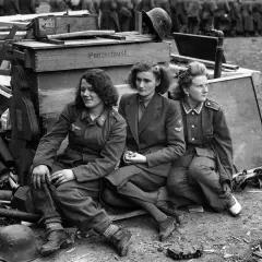 Trzy kobiety w mundurach siedzą obok skrzyń z napisem "Panzerfaust". To poruszająca fotografia wojenna.