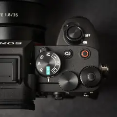 Zbliżenie na górny panel aparatu Sony A7 IV, gotowego na premierę. Widoczny obiektyw FE 1.8/35 i pokrętło trybów.