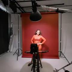 Modelka w pomarańczowej sukience pozuje na tle czerwonego tła. To profesjonalna fotografia studyjna z oświetleniem i sprzętem.