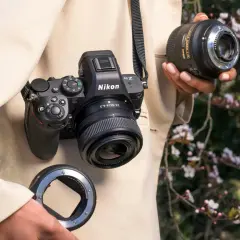 Osoba trzyma aparat Nikon Z5II z obiektywem i adapterem. W tle kwitnące drzewa.