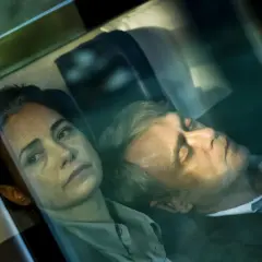 Kobieta patrzy wprost, mężczyzna śpi obok. Scena z filmu, w stylu **June Newton**.