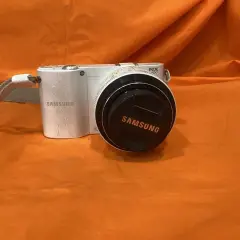 Biały aparat Samsung NX1100 z obiektywem i dekielkiem z logo Samsung, na pomarańczowym tle.