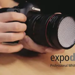 Dłonie trzymają aparat z obiektywem, na którym zamontowano biały dyfuzor. Obok widoczne logo "expodisc Professional White Balance".