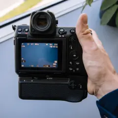 Dłoń trzyma aparat Nikon Z8, testując jego funkcje. Na ekranie widać ustawienia, a w tle rozmyty czerwony samochód.