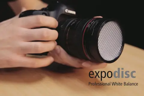 Dłonie trzymają aparat z obiektywem, na którym zamontowano biały dyfuzor. Obok widoczne logo "expodisc Professional White Balance".
