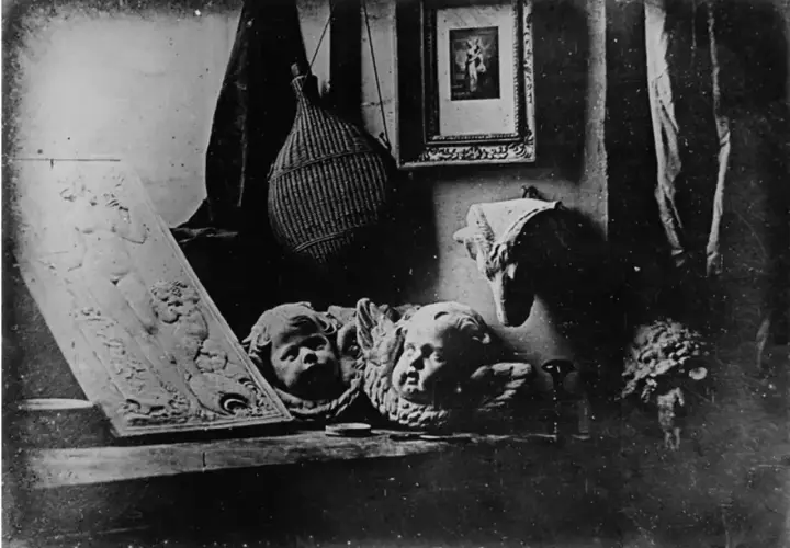 Studiowe ujęcie z początków fotografii: płaskorzeźba z postacią, dwa aniołki, pleciona butla i obrazek.