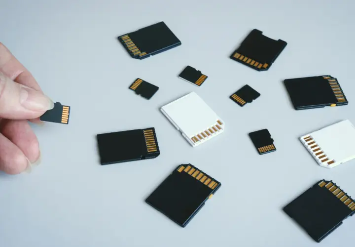 Różne rodzaje kart pamięci: microSD, SD i adaptery. Dłoń trzyma małą kartę microSD, obok leżą inne karty.