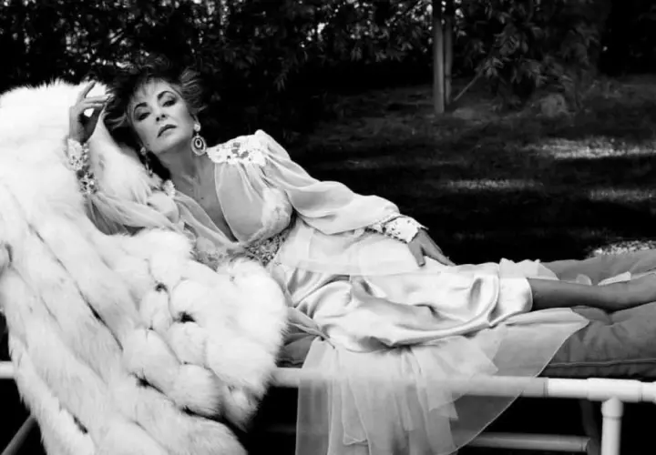 Kobieta w białym futrze i jedwabnej sukni, w stylu Helmut Newton, leży na leżance w ogrodzie.