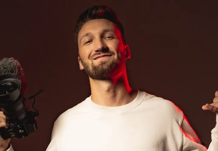 Marcin Kuciński, twórca wideo, z uśmiechem prezentuje swój sprzęt: aparat z mikrofonem i daje kciuk w górę.