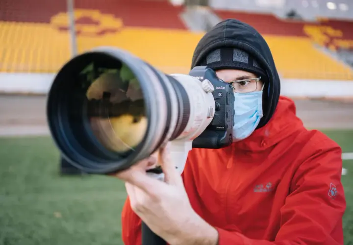Fotograf sportowy w masce i czerwonej kurtce z aparatem Canon EOS-1D X Mark III czeka na akcję na tle trybun stadionu.