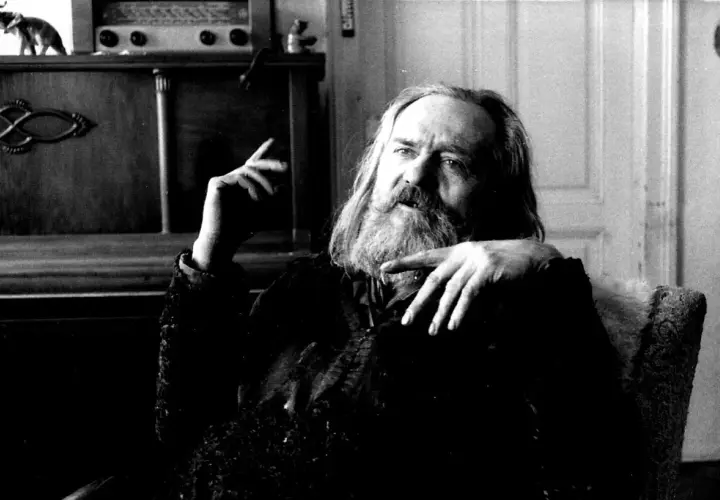 Miroslav Tichý, zamyślony, z długą brodą, siedzi w fotelu. W tle widać mebel z radiem i figurką.