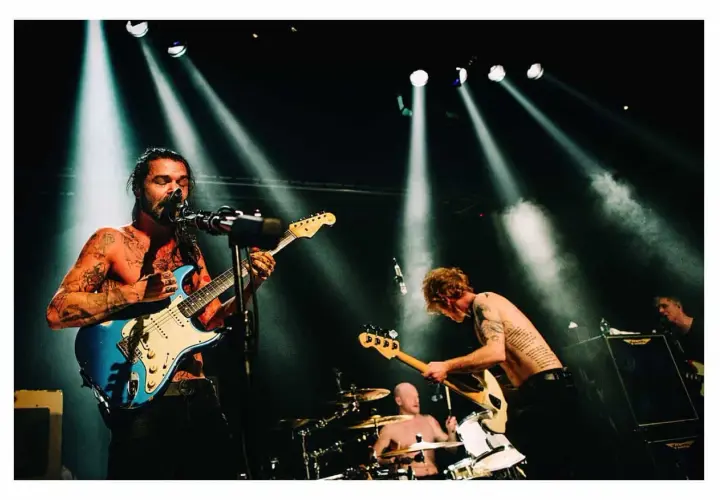Energetyczna fotografia koncertowa: wokalista z tatuażami gra na niebieskiej gitarze, basista pochyla się nad swoim instrumentem, a perkusista wybija rytm.