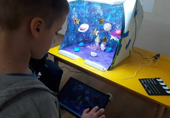 Chłopiec tworzy kosmiczną animację poklatkową. Na stole stoi diorama z rakietą, astronautą i planetami, a obok tablet z podglądem.