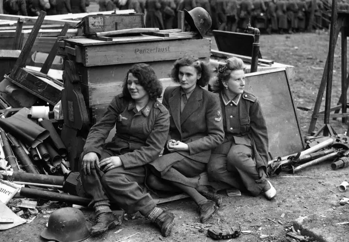 Trzy kobiety w mundurach siedzą obok skrzyń z napisem "Panzerfaust". To poruszająca fotografia wojenna.