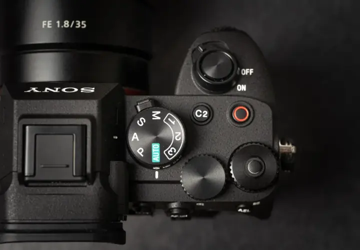 Zbliżenie na górny panel aparatu Sony A7 IV, gotowego na premierę. Widoczny obiektyw FE 1.8/35 i pokrętło trybów.