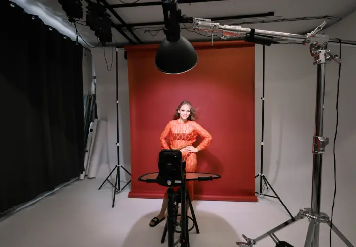 Modelka w pomarańczowej sukience pozuje na tle czerwonego tła. To profesjonalna fotografia studyjna z oświetleniem i sprzętem.