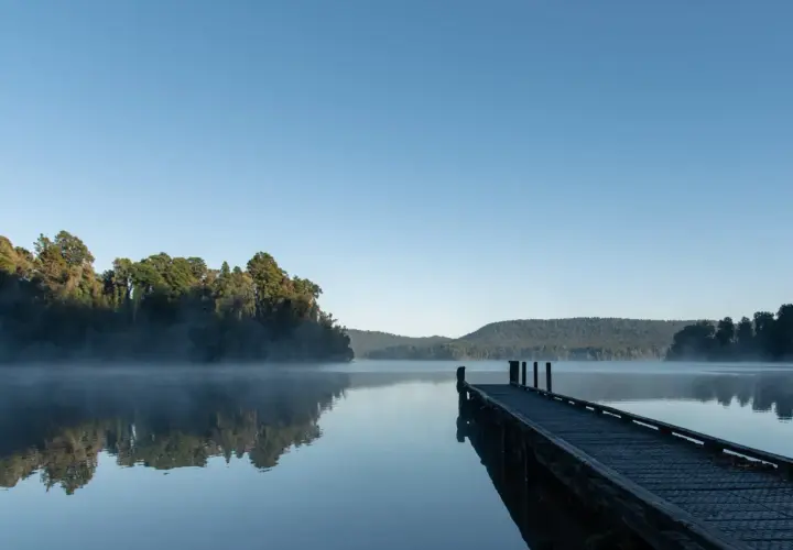 Spokojny wschód słońca nad jeziorem, mgła unosi się nad wodą, a drewniane molo zaprasza do spaceru.