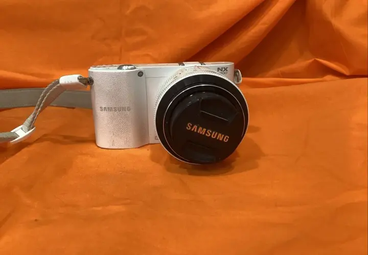 Biały aparat Samsung NX1100 z obiektywem i dekielkiem z logo Samsung, na pomarańczowym tle.
