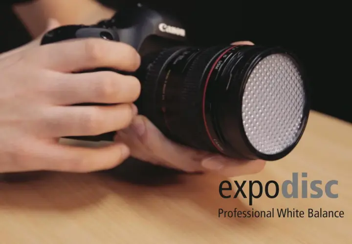 Dłonie trzymają aparat z obiektywem, na którym zamontowano biały dyfuzor. Obok widoczne logo "expodisc Professional White Balance".