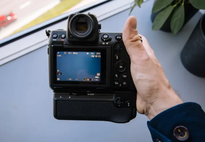 Dłoń trzyma aparat Nikon Z8, testując jego funkcje. Na ekranie widać ustawienia, a w tle rozmyty czerwony samochód.