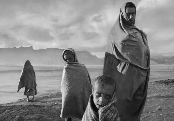 Dzieci i dorosły owinięci w tkaniny, w surowym krajobrazie. Wzrok przyciąga spojrzenie jednego z nich, uchwycone przez Sebastiao Salgado.