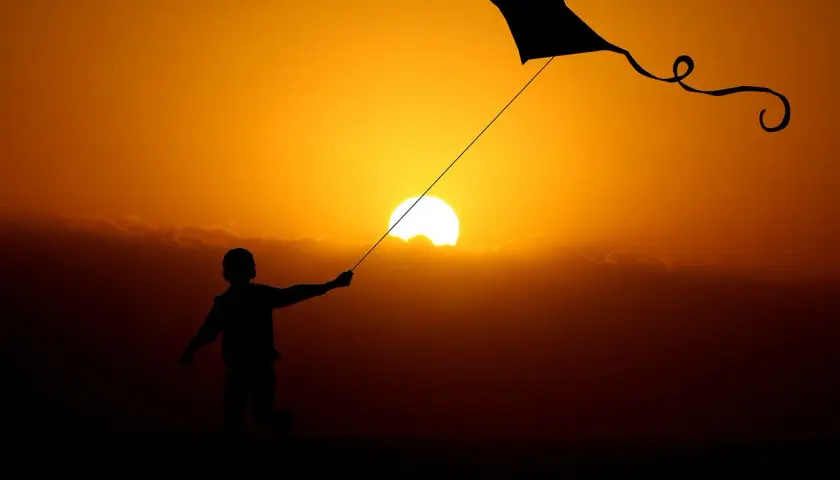 Dziecko puszcza latawiec na tle zachodzącego słońca. Ciepłe światło w fotografii podkreśla sylwetki i tworzy magiczną atmosferę.
