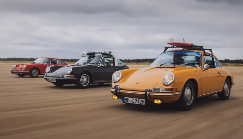 Trzy klasyczne Porsche jadące po plaży. Kompozycja dynamiczna fotografia z deskami surfingowymi na dachu.
