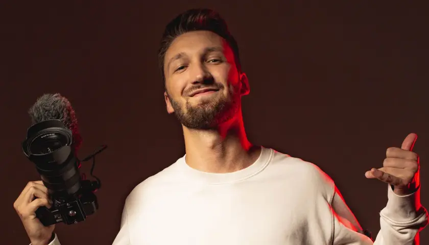 Marcin Kuciński, twórca wideo, z uśmiechem prezentuje swój sprzęt: aparat z mikrofonem i daje kciuk w górę.