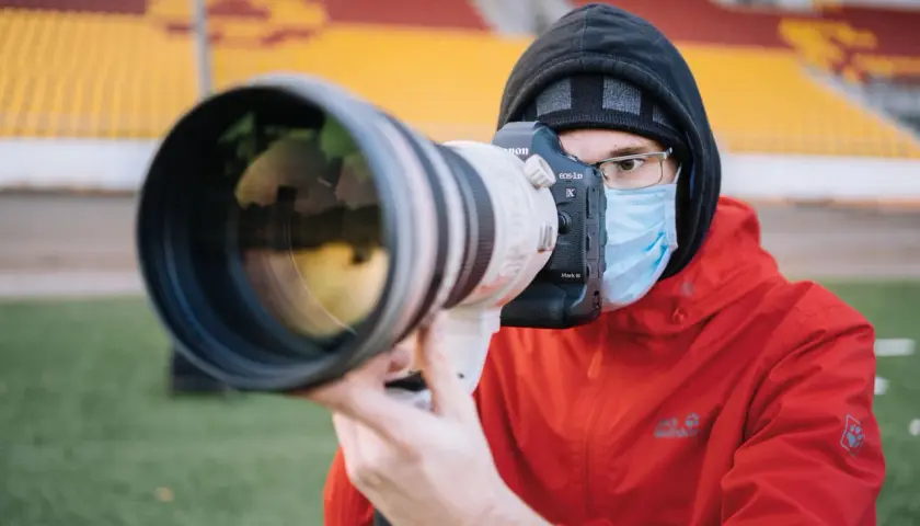 Fotograf sportowy w masce i czerwonej kurtce z aparatem Canon EOS-1D X Mark III czeka na akcję na tle trybun stadionu.