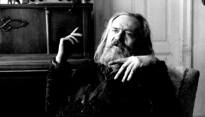 Miroslav Tichý, zamyślony, z długą brodą, siedzi w fotelu. W tle widać mebel z radiem i figurką.