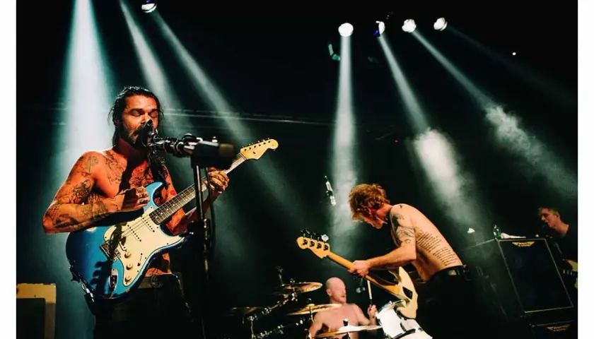 Energetyczna fotografia koncertowa: wokalista z tatuażami gra na niebieskiej gitarze, basista pochyla się nad swoim instrumentem, a perkusista wybija rytm.