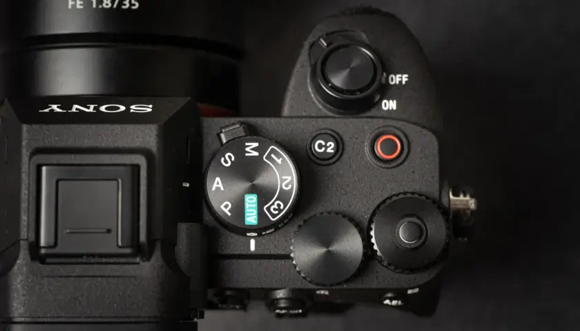Zbliżenie na górny panel aparatu Sony A7 IV, gotowego na premierę. Widoczny obiektyw FE 1.8/35 i pokrętło trybów.
