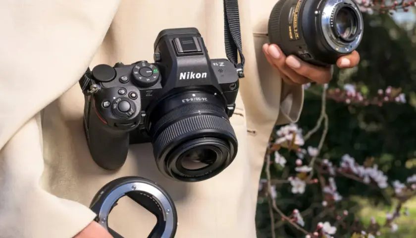 Osoba trzyma aparat Nikon Z5II z obiektywem i adapterem. W tle kwitnące drzewa.