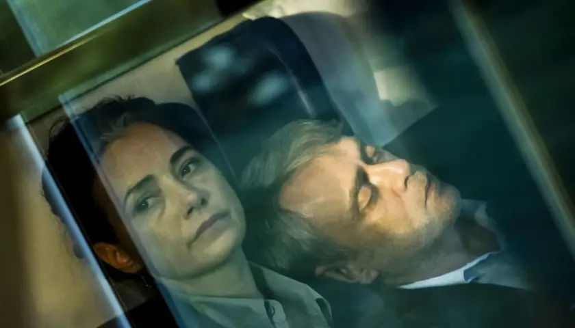 Kobieta patrzy wprost, mężczyzna śpi obok. Scena z filmu, w stylu **June Newton**.