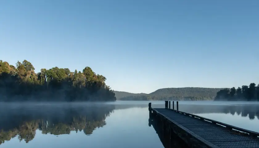 Spokojny wschód słońca nad jeziorem, mgła unosi się nad wodą, a drewniane molo zaprasza do spaceru.