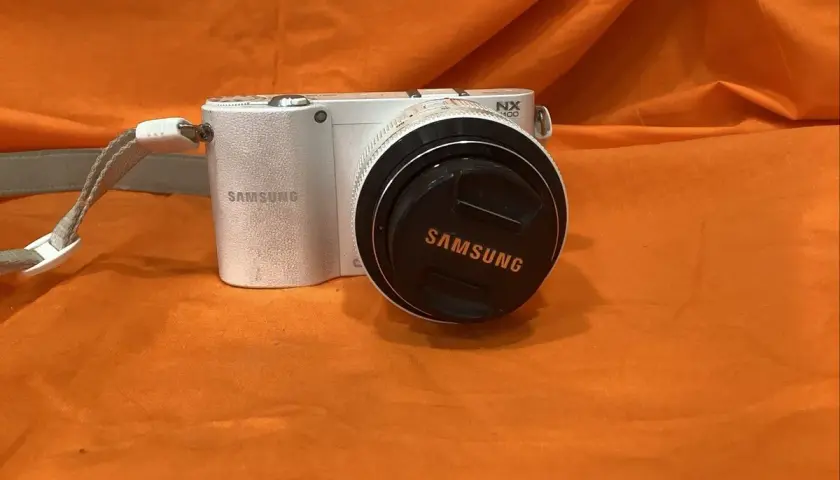 Biały aparat Samsung NX1100 z obiektywem i dekielkiem z logo Samsung, na pomarańczowym tle.