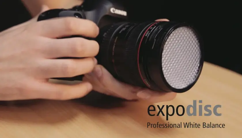 Dłonie trzymają aparat z obiektywem, na którym zamontowano biały dyfuzor. Obok widoczne logo "expodisc Professional White Balance".