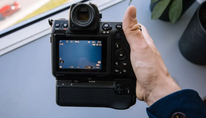 Dłoń trzyma aparat Nikon Z8, testując jego funkcje. Na ekranie widać ustawienia, a w tle rozmyty czerwony samochód.