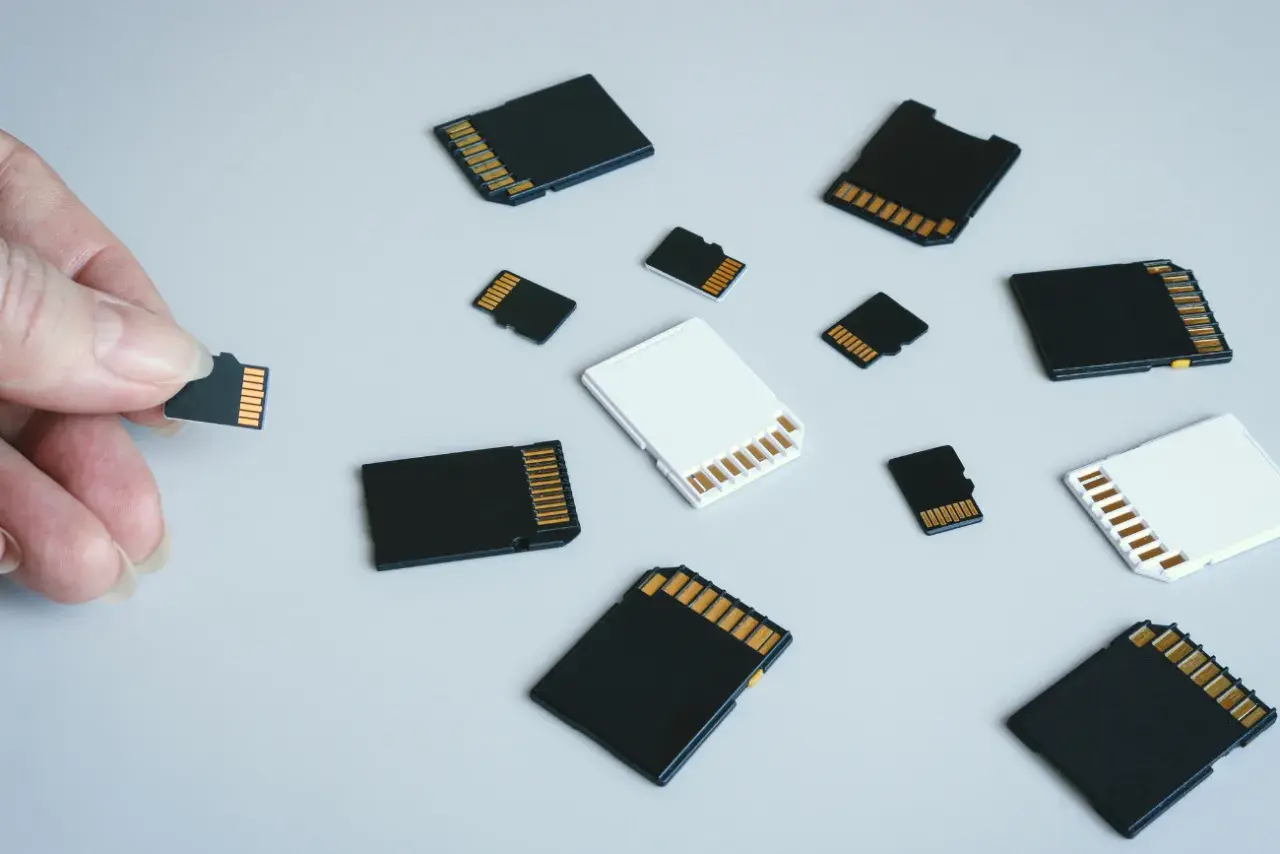 Różne rodzaje kart pamięci: microSD, SD i adaptery. Dłoń trzyma małą kartę microSD, obok leżą inne karty.
