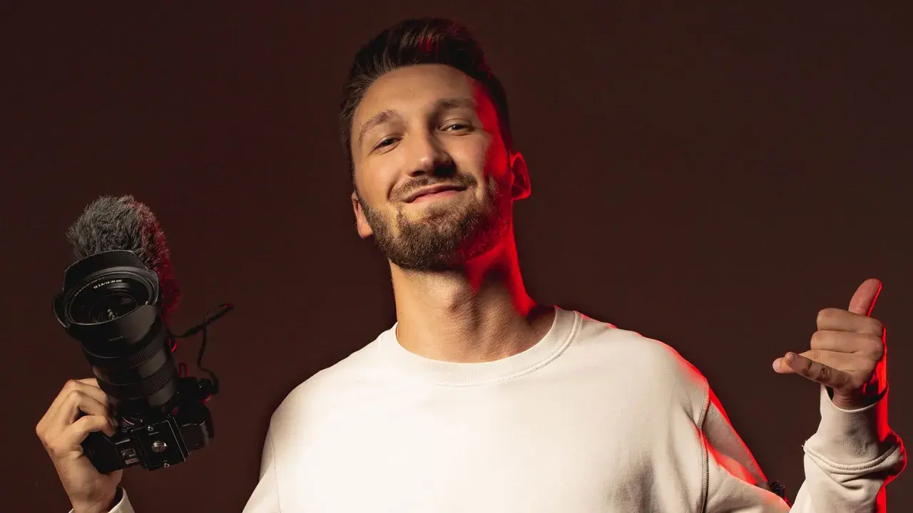 Marcin Kuciński, twórca wideo, z uśmiechem prezentuje swój sprzęt: aparat z mikrofonem i daje kciuk w górę.