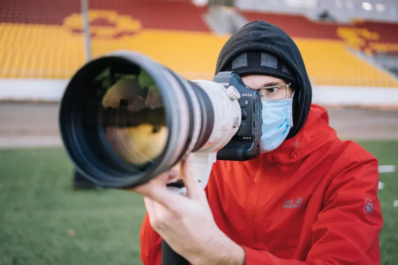 Fotograf sportowy w masce i czerwonej kurtce z aparatem Canon EOS-1D X Mark III czeka na akcję na tle trybun stadionu.