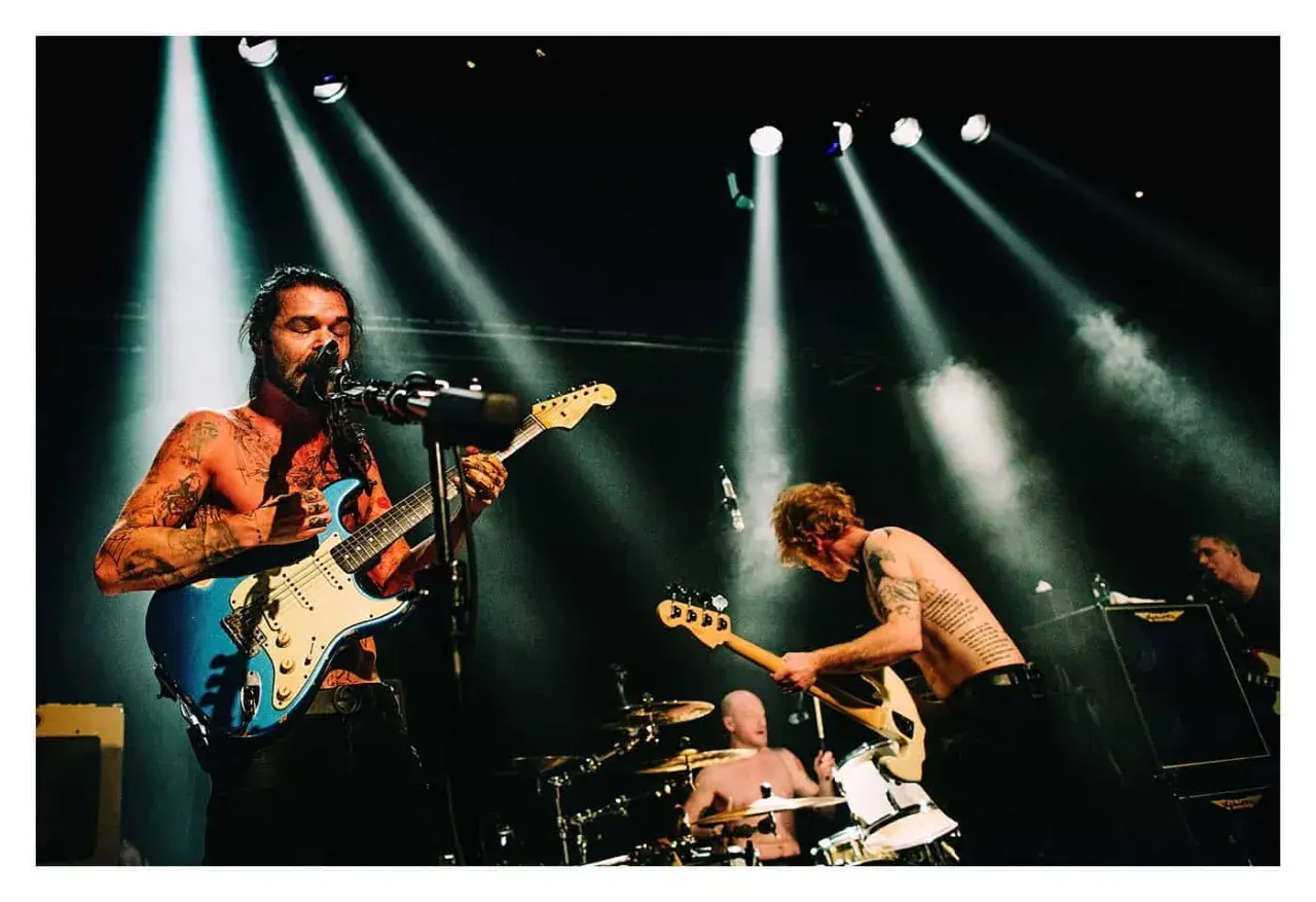 Energetyczna fotografia koncertowa: wokalista z tatuażami gra na niebieskiej gitarze, basista pochyla się nad swoim instrumentem, a perkusista wybija rytm.
